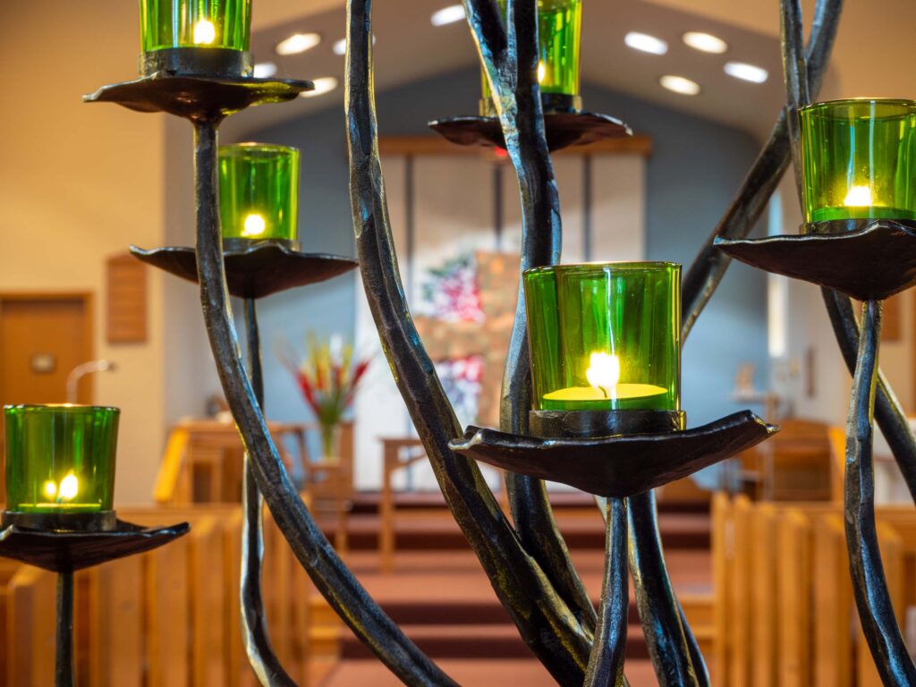 A close up of green votive glasses with lit candles inside of them on the metal branches of a prayer tree. In the background is a view of a church sanctuary.