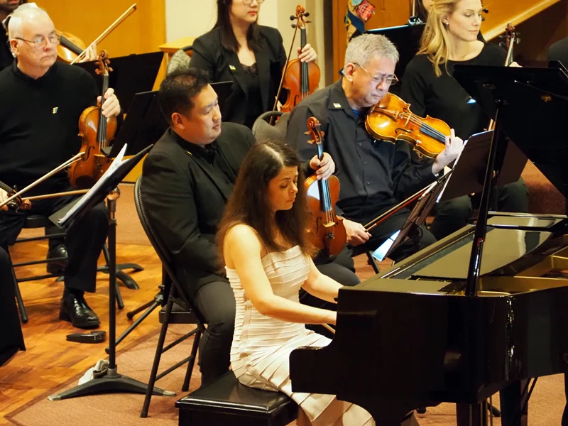 Ukrainian pianist, Anna Sagalova, playing a baby grand piano, surrounded by violinists during a concert.