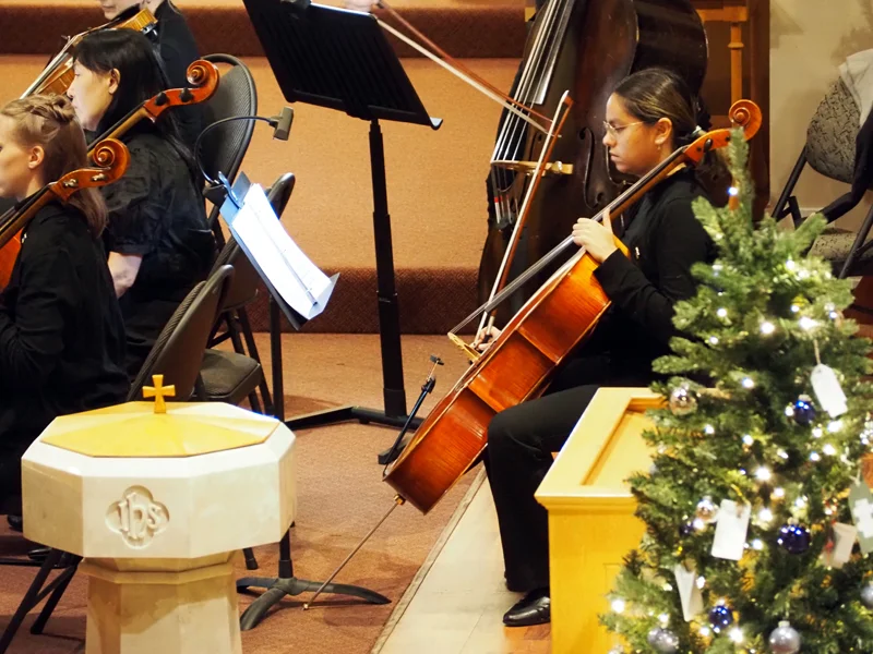 String musician seated with her instrument with the top of a Christmas tree and baptismal font in the foreground.